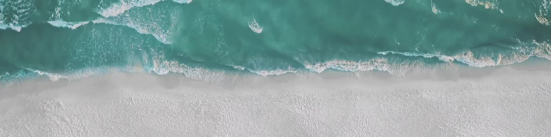 Blue-turquoise surf meets an open stretch of pale coastal sand.